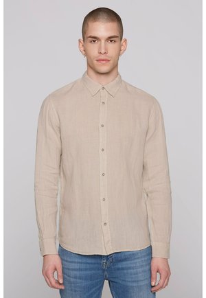 Mann mit kurzen Haaren und Ohrringen, der ein beige-farbenes, langärmliges Hemd mit Knöpfen und blaue Jeans trägt, steht vor einem einfarbigen Hintergrund.