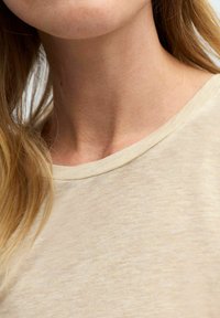 Lys beige t-shirt med et blødt, tekstureret stof og en rund halsudskæring. Synlige syninger langs kraven og skulderområdet.
