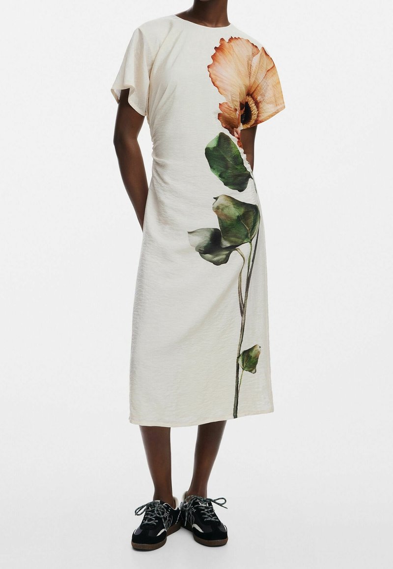 Personne portant une robe midi blanche à manches courtes avec un grand motif de fleur orange et de feuilles vertes à l'avant, associée à des baskets noires.