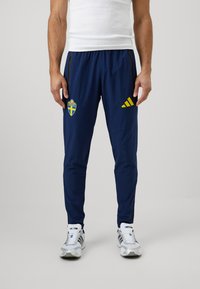Homem com calças de desporto azul-marinho com o emblema da seleção sueca de futebol amarelo e o logótipo da Adidas, combinado com ténis brancos da Adidas.
