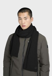Sort ribbet strikscarf båret over en brun jakke, med en glat tekstur og rektangulær form, prydet med et logo i enden.