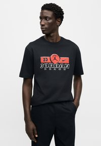 Μαύρο βαμβακερό t-shirt με στρογγυλή λαιμόκοψη, με γραφικό τύπωμα κόκκινου και άσπρου χρώματος που γράφει "JORDAN" και διαθέτει λογότυπο, κοντά μανίκια και άνετη εφαρμογή.