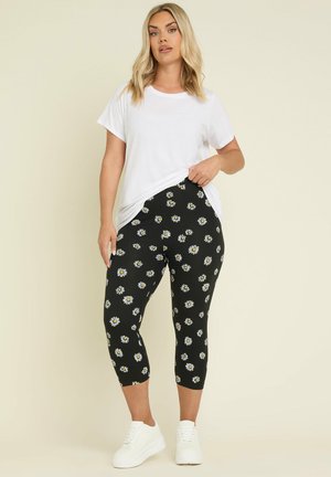 Leggings negras con estampado de margaritas blancas y amarillas, de longitud corta, combinadas con una camiseta blanca de corte holgado y zapatillas blancas.
