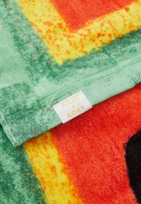 Roxy strandlaken met levendige kleuren - rood, geel, groen en zwart. Zachte textuur, abstract patroon en een wit label met "ATLAS × ROXY."