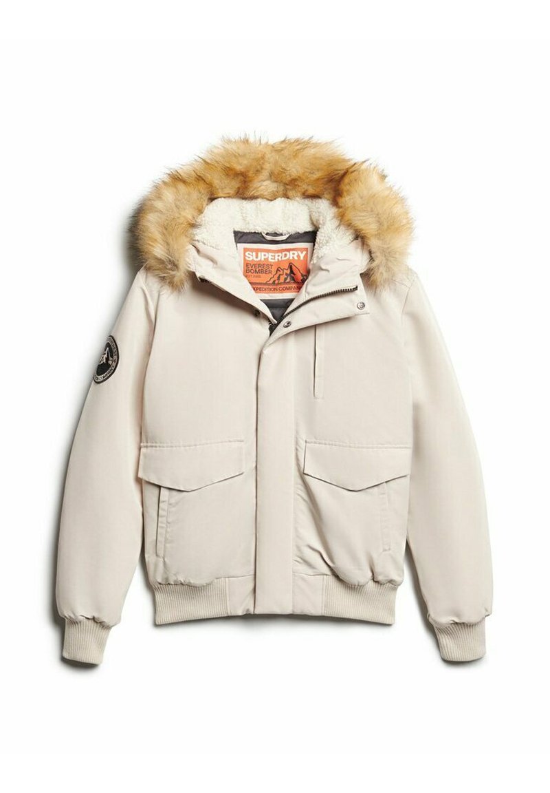 Beige Bomberjacke mit einer Kapuze aus Kunstfell, zwei Fronttaschen, Rippbündchen und einem orangefarbenen Etikett im Innenfutter. Glatte Außenoberfläche.