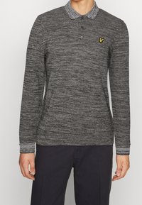 Homme portant un polo à manches longues gris foncé avec un logo d'oiseau jaune sur la poitrine gauche et un pantalon noir.