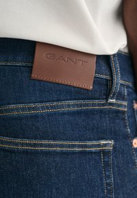 Jeans in denim blu scuro con cuciture gialle a contrasto e una etichetta in pelle marrone con testo in rilievo, posizionata vicino alla vita.