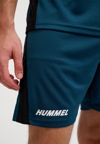 Teal sportshorts van ademend materiaal, met zwarte zijpanelen, stikdetails en een wit "HUMMEL" logo aan de onderrand.