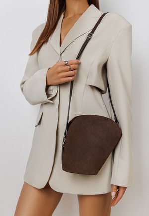 Rjava semišan crossbody torba s ukrivljeno zasnovo, ki ima temen trak in srebrno okovje. Nosimo jo z bež oversize blejzerjem.
