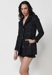 Sacou din tweed negru cu model în carouri albe, margini desfăcute, colier cu decupaj și nasturi în față, asortat cu pantaloni scurți coordonați.