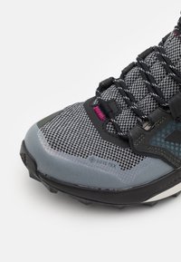 Chaussure de course grise avec une tige en mesh, un bout renforcé et une semelle extérieure en caoutchouc noir. Présente une étiquette GORE-TEX et des accents réfléchissants.