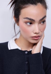 Cardigan bleu marine foncé avec fermeture à boutons et poignets côtelés, porté sur une chemise blanche à col. Peau lisse avec un maquillage naturel et des traits doux.