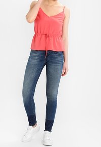 Top peplum de coral con tirantes ajustables, combinado con jeans skinny de lavado oscuro y zapatillas blancas. Tejido suave y detalle de cintura ceñida.