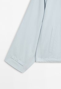 Manica di giacca azzurro chiaro con dettaglio di cinturino con bottone vicino al polsino e tasca laterale verticale sulla sezione del corpo.
