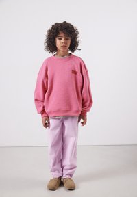 Enfant aux cheveux bouclés portant un sweat-shirt rose oversize, un pantalon violet clair et des chaussures marron sans lacets, se tenant devant un fond uni.