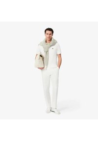 Bílá polo košile s logem Lacoste, šedý pletený šátek, béžová taška, bílé kalhoty a bílo-zelené tenisky, stojící na jednobodovém pozadí.