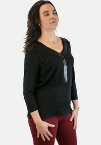 Top negro de manga larga con escote en V, que presenta un detalle texturizado y un cierre con cremallera adornado con lentejuelas. Combinado con pantalones burdeos.