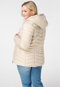 Veste rembourrée beige avec capuche, dotée de lignes cousues verticalement, d'une fermeture éclair et de détails côtelés au niveau du col.