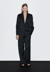 Czarny garnitur szyty na miarę z głębokim dekoltem w kształcie V i szerokimi spodniami, z dwoma frontowymi guzikami i wyraźnie zarysowanymi ramionami. Gładka, matowa faktura.