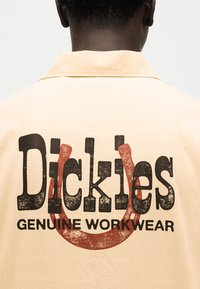 Πίσω όψη ενός ατόμου που φορά μπλούζα μπεζ με φθαρμένο μαύρο κείμενο "Dickies Genuine Workwear" και ένα κόκκινο γραφικό πετάλου.