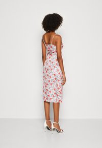 Robe mi-longue fleurie à fines bretelles, avec un fond rose clair, des motifs floraux rouges et bleus, et une silhouette ajustée.