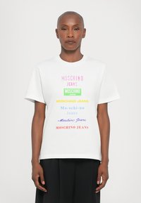 White t-shirt featuring multi-colored "MOSCHINO JEANS" text in varying fonts, short sleeves, and a crew neck. Smooth fabric texture.