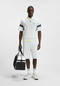 Hvid strikket polo med korte ærmer og lynlås krave, grå accenter, lysegrå shorts og en sort og hvid duffeltaske. Hvide sneakers og logo-strømper.