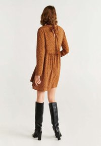 Robe à motifs marron avec manches longues, taille cintrée et un lien au col, associée à des bottes noires montantes jusqu'aux genoux. Texture lisse.