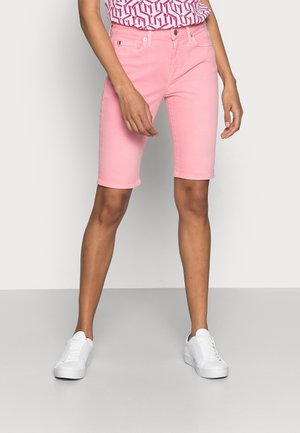 Person iført knælange pink shorts, mønstret pink og hvid top og hvide sneakers, stående mod en ensfarvet baggrund.