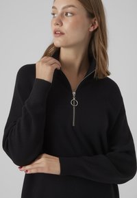 Maglione nero a collo con zip a metà, polsini a coste e finitura testurizzata. Presenta un accento di zip a forma circolare.