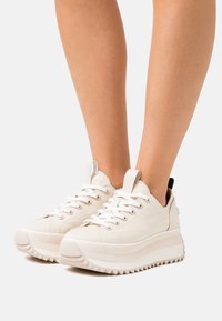 Weiße Leinensneaker mit einer massiven, strukturierten Sohle, runden Schnürsenkeln und einer Schlaufe an der Ferse, vor einem hellen Hintergrund.