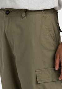 Cargo byxor i olivgrön tyg, med en smal passform, texturerat material och en sidoficka med lock. Stylistiskt med kontrasterande sömmar.