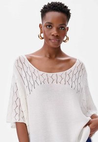 Witte gebreide top met een losse pasvorm, brede ronde halslijn en een textuurpatroon met opengewerkte details. Korte mouwen en een zachte textuur.