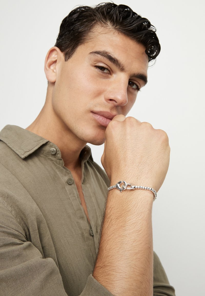 Pulsera de plata con diseño de cierre, con eslabones redondeados y texturizados, llevada en una muñeca. La persona tiene el cabello corto, oscuro y peinado.