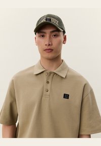 Beige polo-shirt med klassisk krage, tre-knapps placket og en sort logo-patch på brystet. Bæres med en grøn caps.