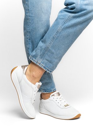 Persoon draagt witte sneakers met metallic accenten en lichtblauwe jeans, poserend met gekruiste benen tegen een witte achtergrond.