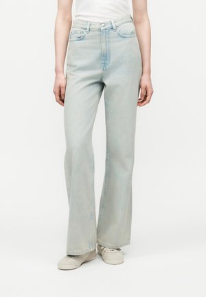 Person trägt hellblaue, hoch taillierte, weit geschnittene Jeans und weiße Turnschuhe, steht vor einem schlichten weißen Hintergrund.