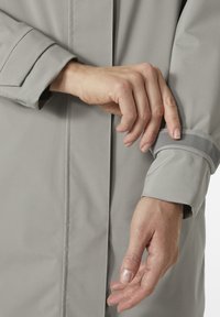 Světle šedá nepromokavá bunda s přehnutým manžetou a reflexním proužkem, hladkou texturou a čistými liniemi. Detail ruky, která upravuje manžetu.