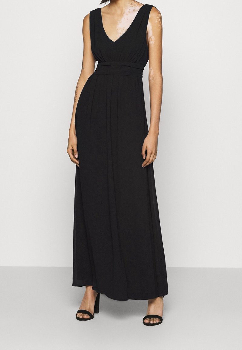 Femme portant une robe maxi noire sans manches avec un décolleté en V et une taille cintrée, associée à des talons hauts noirs à bouts ouverts.