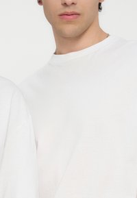 Giovane persona indossa una maglietta bianca semplice con scollo rotondo, mostrando la parte bassa del viso e il collo con carnagione chiara e una collana a catena d'argento.