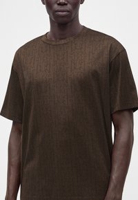 Σκούρα καφέ κοντομάνικο T-shirt με διακριτικό τονικό μοτίβο με μονόγραμμα, φορεμένο από άτομο με σκούρο δέρμα σε απλό ανοιχτό φόντο.
