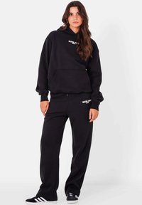 Zwarte katoenen hoodie met grote voorzak en logo-opdruk, gecombineerd met bijpassende zwarte sweatpants. Beide hebben een losse pasvorm en een boordontwerp.