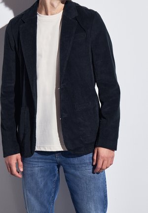Man draagt een zwart corduroy blazer, wit T-shirt en blauwe spijkerbroek tegen een effen lichte achtergrond.