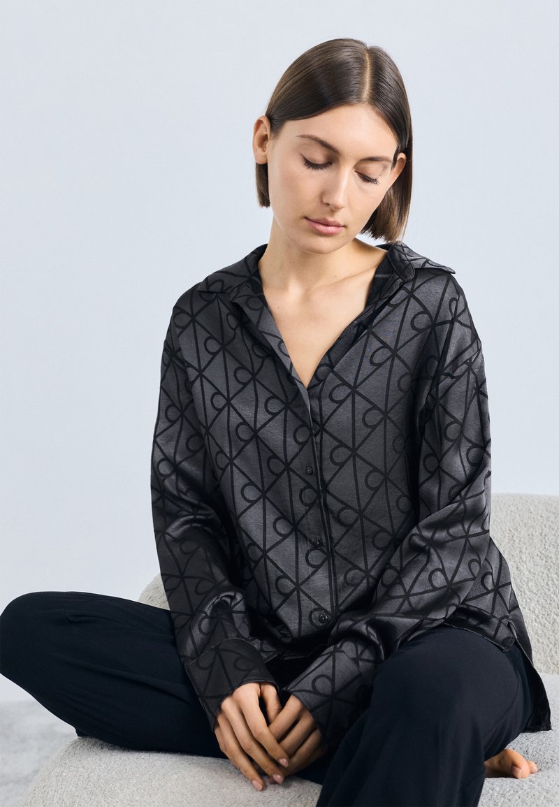 Cămașă satinată gri închis cu un model geometric negru, mâneci lungi și guler, asortată cu pantaloni negri. Textură netedă și design cu nasturi pe față.