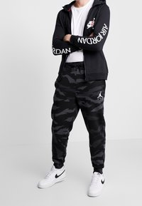 Černá mikina na zip s logem "Nike", doplněná černými maskáčovými tepláky s bílým logem "Jordan" a bílými teniskami Nike.