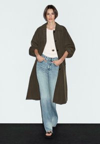 Cappotto lungo verde oliva scuro, top bianco, jeans azzurri chiari, sandali neri. Modella posa con le mani in tasca, vestibilità ampia, design minimalista.