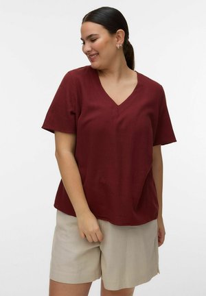 Frau lächelt, trägt ein burgunderrotes V-Ausschnitt-Shirt mit kurzen Ärmeln und beige knielange Shorts, steht vor einem schlichten weißen Hintergrund.