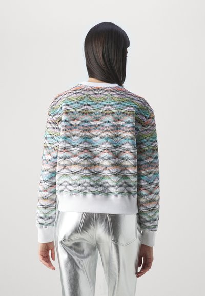 Απαλή κοντή μπλούζα με υφή σε πολύχρωμο σχέδιο ζιγκ-ζαγκ, με λευκή πλεκτή λαιμόκοψη και μανίκια, συνδυασμένη με γυαλιστερούς ασημένιους παντελόνες.