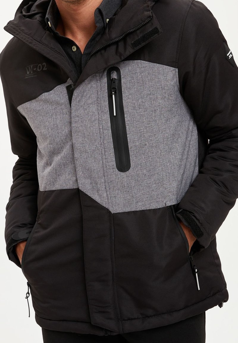 Černá a šedá prošívaná bunda s kapsou na zip, strukturovaným předním dílem, nastavitelnou kapucí a kontrastními barevnými bloky. Obsahuje boční kapsy.