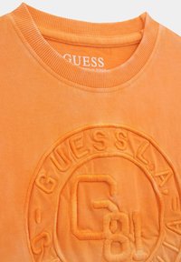 Oranža kokvilnas sporta krekls ar apaļu kaklu un teksturētu logo izšuvumu ar "GUESS" un "L.A." detaļām. Maiga auduma apdare.